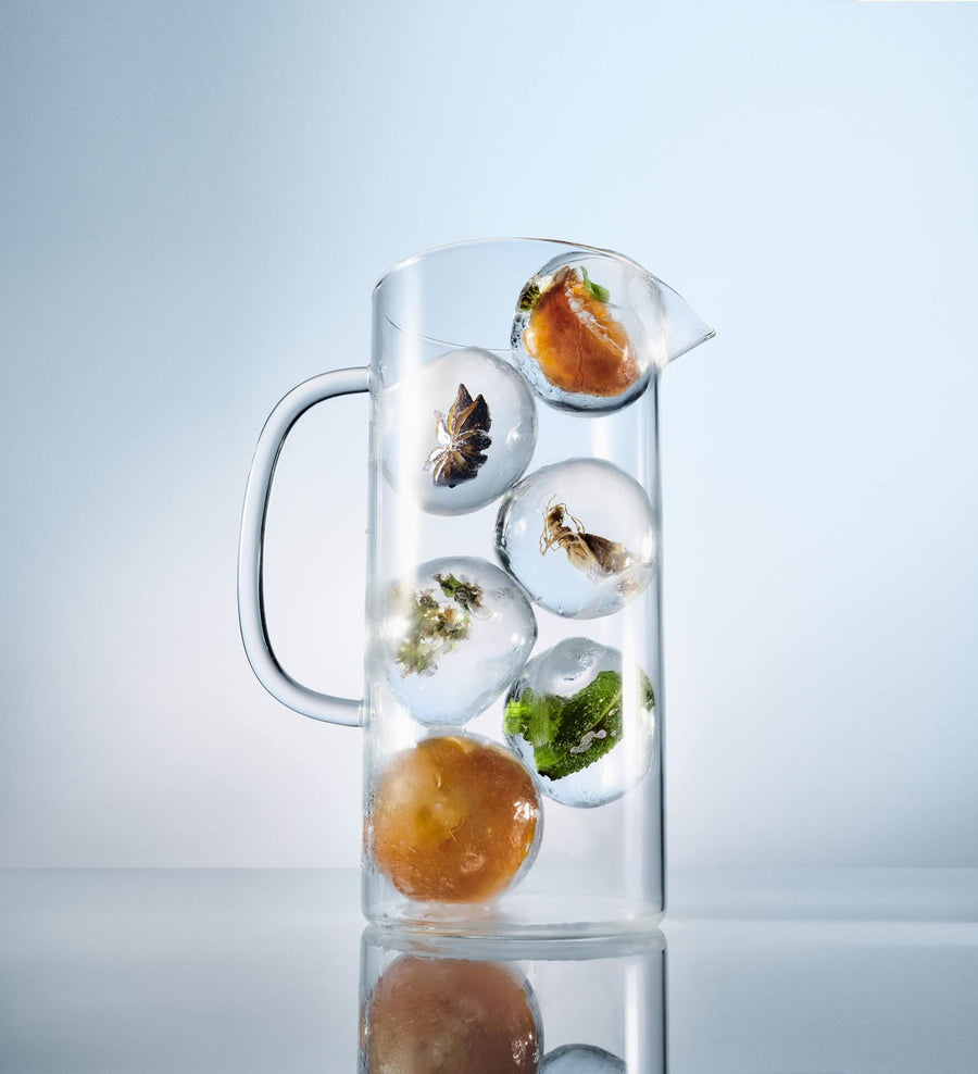 Glass jug, pitcher containing round ice cubes filled with apricot, lemon balm, anise, panax ginseng and za'atar, on a blue gradient backdrop.